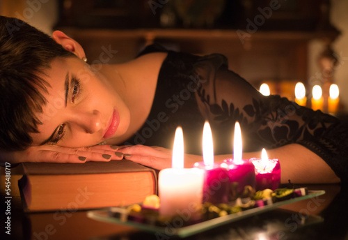Beautiful young woman in old fashioned black dress reading book under candlelight at night