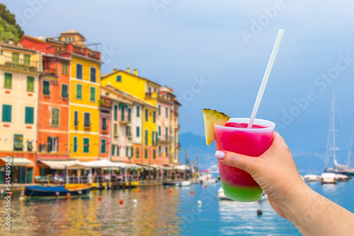 Portofino with colorful houses and villas in little bay harbor with hand holding a glass with cocktail and straw. Liguria, Italy, Europe
