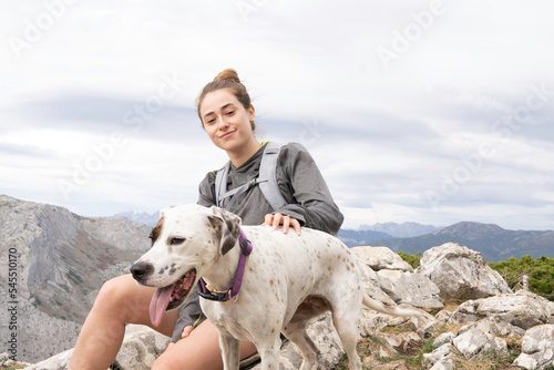 a young smiling white girl dressed in mountain clothes and a gray backpack. Her dog accompanies her on this adventure. In the background you can see the mountain range.
