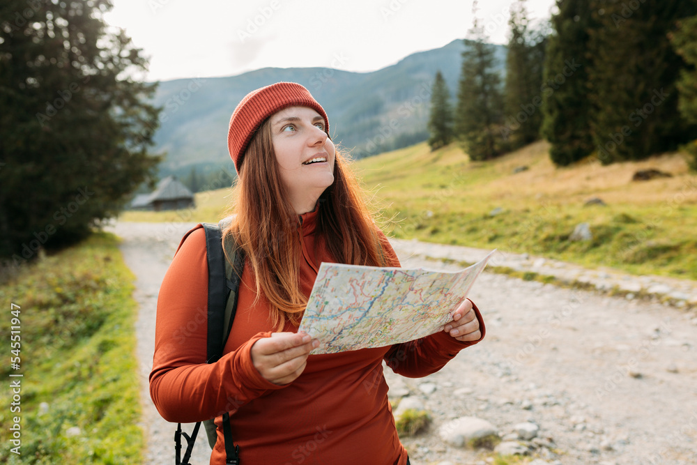 Stylish woman holding paper map relaxing in nature. Travel and active lifestyle concept. Hiking active female lifestyle wearing backpack and red knit hat exercising outdoors.