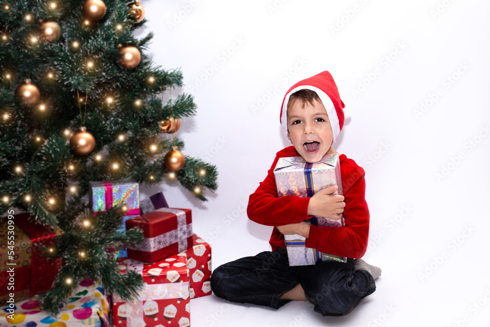 cute boy near the christmas tree holding a gift. happy child on a white background in a red New Year's hat and a red jacket hugs a gift. cute boy near the christmas tree holding a gift. happy child on a white background in a red New Year's hat and a red jacket hugs a gift.