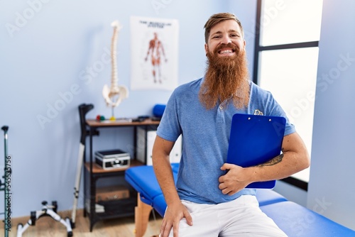 Young redhead man wearing physiotherapist uniform holding clipboard at physiotherapy clinic
