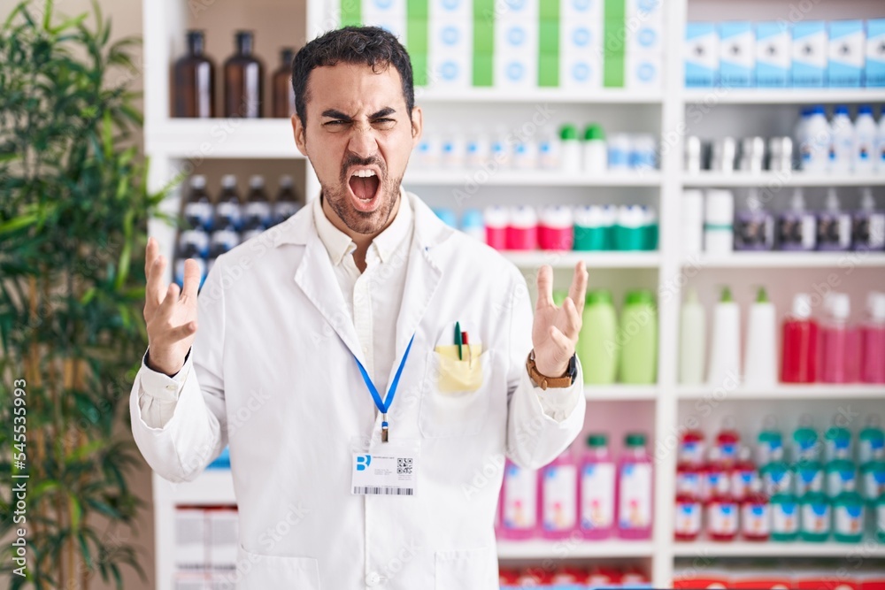 Handsome hispanic man working at pharmacy drugstore crazy and mad shouting and yelling with aggressive expression and arms raised. frustration concept.