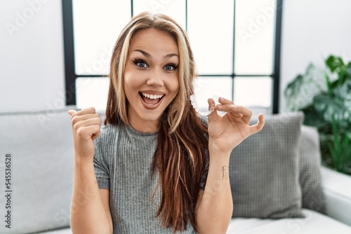 Young caucasian woman holding invisible aligner orthodontic pointing thumb up to the side smiling happy with open mouth