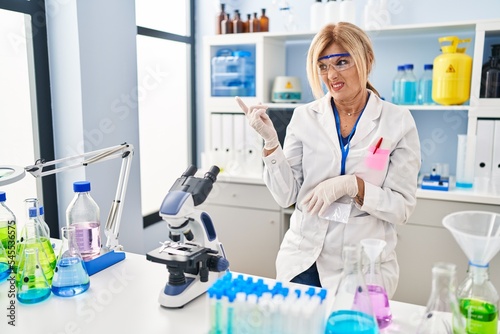 Middle age blonde woman working at scientist laboratory pointing aside worried and nervous with forefinger  concerned and surprised expression