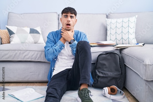 Young hispanic man sitting on the floor studying for university shouting suffocate because painful strangle. health problem. asphyxiate and suicide concept.