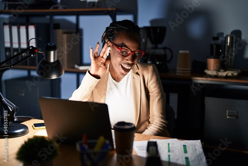 Beautiful black woman working at the office at night smiling with hand over ear listening an hearing to rumor or gossip. deafness concept.