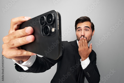 Man businessman in suit takes selfies on phone posing in front of smartphone camera with smile with teeth happy win on gray background close-up face wide camera angle