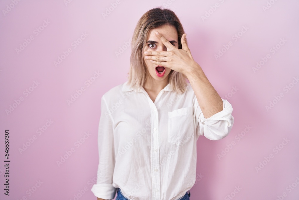 Young beautiful woman standing over pink background peeking in shock covering face and eyes with hand, looking through fingers with embarrassed expression. Young beautiful woman standing over pink background peeking in shock covering face and eyes with hand, looking through fingers with embarrassed expression.