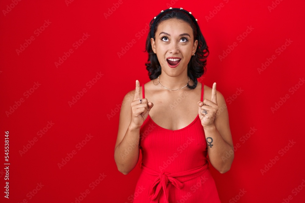 Young hispanic woman standing over red background amazed and surprised looking up and pointing with fingers and raised arms. Young hispanic woman standing over red background amazed and surprised looking up and pointing with fingers and raised arms.