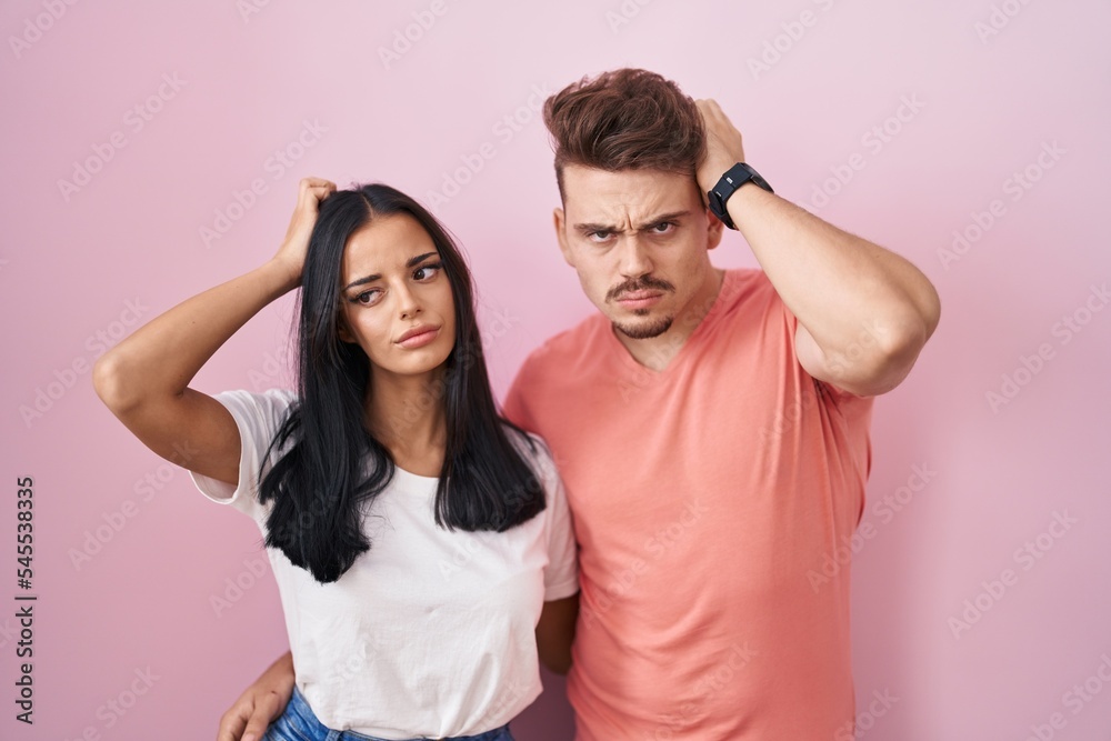 Obraz premium Young hispanic couple standing over pink background confuse and wondering about question. uncertain with doubt, thinking with hand on head. pensive concept.