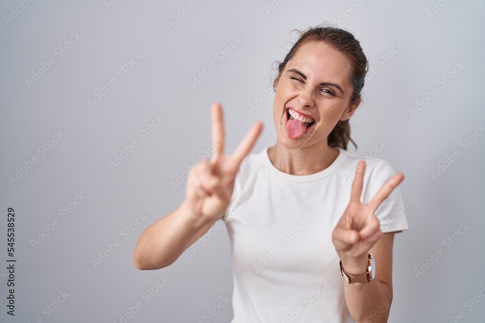 Beautiful brunette woman standing over isolated background smiling with tongue out showing fingers of both hands doing victory sign. number two.