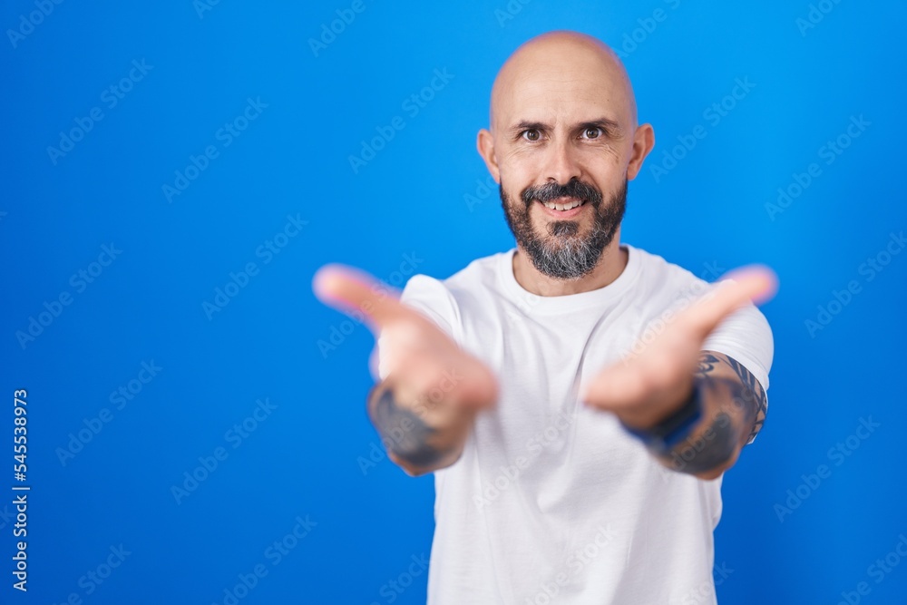 Hispanic man with tattoos standing over blue background smiling with hands palms together receiving or giving gesture. hold and protection Hispanic man with tattoos standing over blue background smiling with hands palms together receiving or giving gesture. hold and protection
