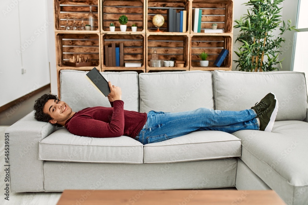 Young hispanic man reading book lying on the sofa at home. Young hispanic man reading book lying on the sofa at home.