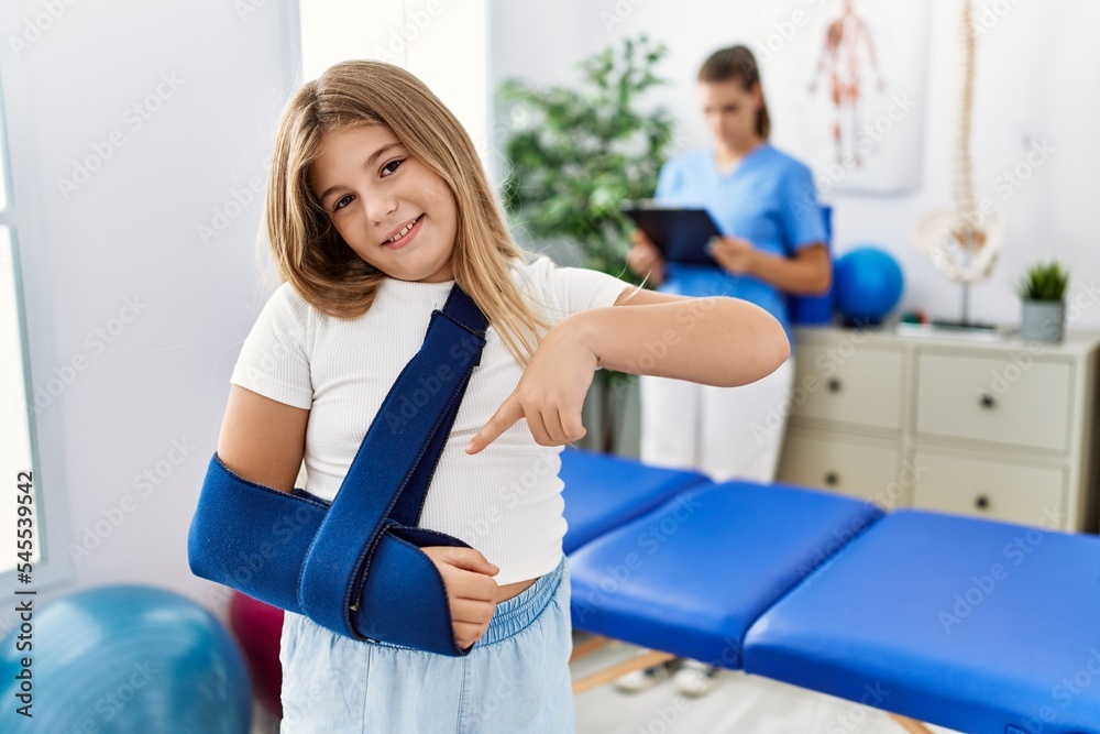 Blonde little girl wearing arm on sling at rehabilitation clinic smiling happy pointing with hand and finger