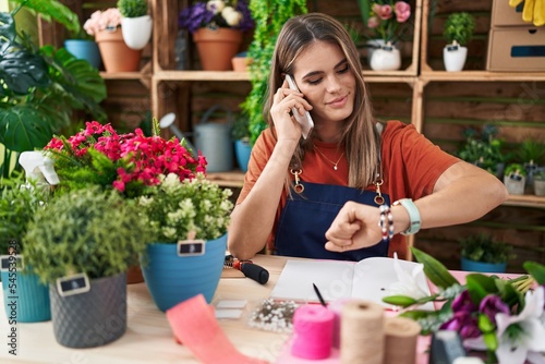 Young beautiful hispanic woman florist talking on smartphone looking watch at flower shop