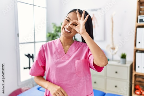 Young hispanic woman working at pain recovery clinic doing ok gesture with hand smiling, eye looking through fingers with happy face.