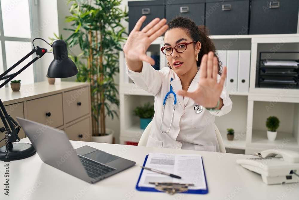 Young hispanic woman wearing doctor uniform and stethoscope doing frame using hands palms and fingers, camera perspective Young hispanic woman wearing doctor uniform and stethoscope doing frame using hands palms and fingers, camera perspective