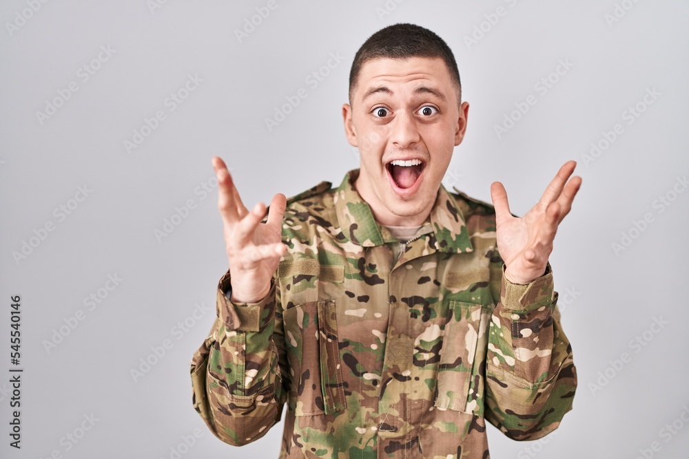 Young man wearing camouflage army uniform celebrating crazy and amazed for success with arms raised and open eyes screaming excited. winner concept Young man wearing camouflage army uniform celebrating crazy and amazed for success with arms raised and open eyes screaming excited. winner concept