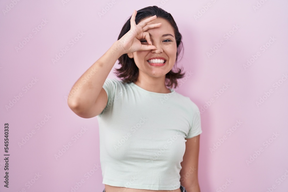 Hispanic young woman standing over pink background doing ok gesture with hand smiling, eye looking through fingers with happy face. Hispanic young woman standing over pink background doing ok gesture with hand smiling, eye looking through fingers with happy face.
