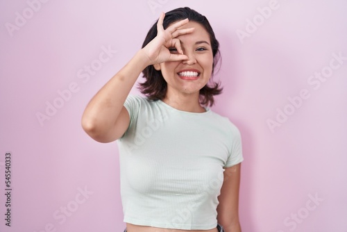 Hispanic young woman standing over pink background doing ok gesture with hand smiling, eye looking through fingers with happy face.