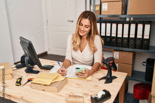 Young blonde woman ecommerce business worker using smartphone and writing on book at office