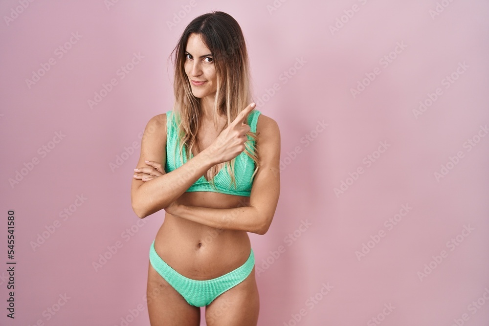 Young hispanic woman wearing bikini over pink background pointing with hand finger to the side showing advertisement, serious and calm face Young hispanic woman wearing bikini over pink background pointing with hand finger to the side showing advertisement, serious and calm face