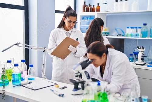Three woman scientists using microscope write on checklist at laboratory