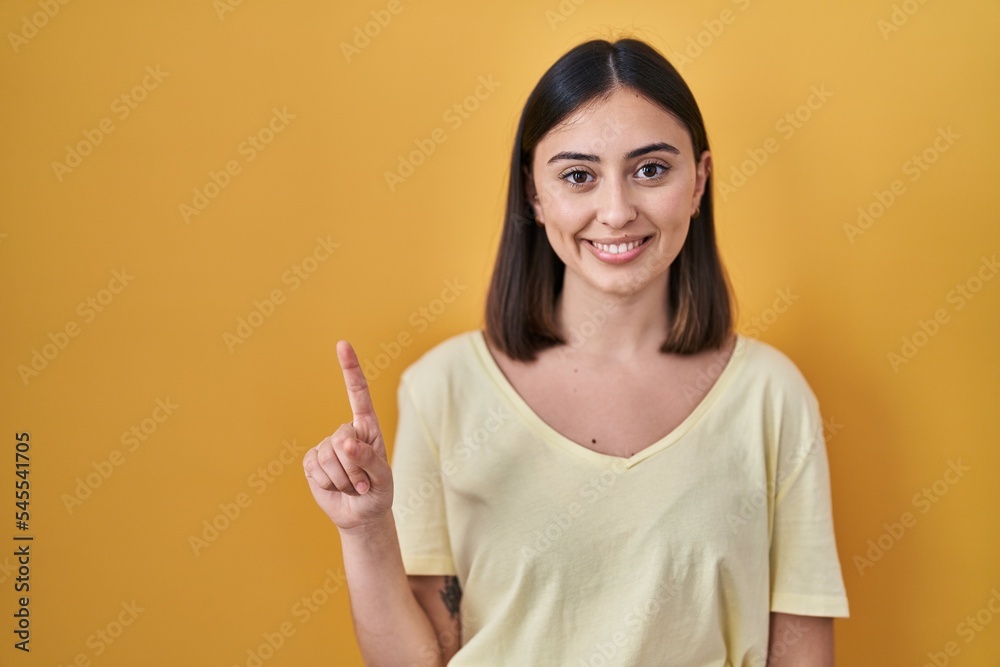 Hispanic girl wearing casual t shirt over yellow background showing and pointing up with finger number one while smiling confident and happy. Hispanic girl wearing casual t shirt over yellow background showing and pointing up with finger number one while smiling confident and happy.