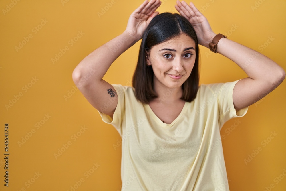Hispanic girl wearing casual t shirt over yellow background doing bunny ears gesture with hands palms looking cynical and skeptical. easter rabbit concept. Hispanic girl wearing casual t shirt over yellow background doing bunny ears gesture with hands palms looking cynical and skeptical. easter rabbit concept.
