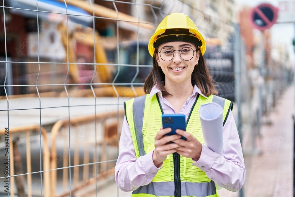 Young hispanic woman architect using smartphone holding blueprints at street Young hispanic woman architect using smartphone holding blueprints at street