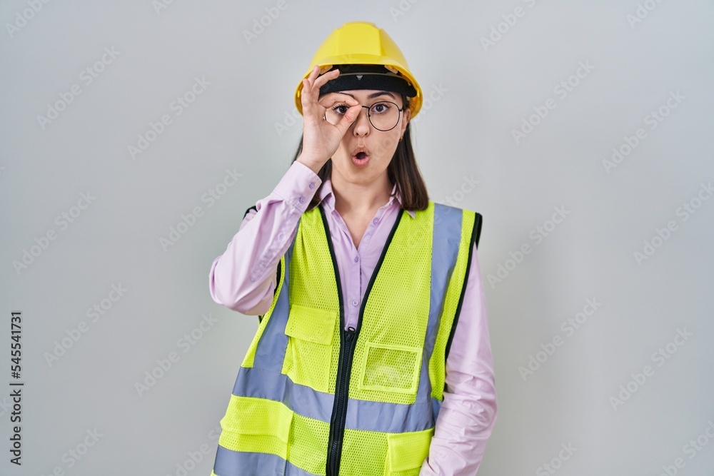Hispanic girl wearing builder uniform and hardhat doing ok gesture shocked with surprised face, eye looking through fingers. unbelieving expression. Hispanic girl wearing builder uniform and hardhat doing ok gesture shocked with surprised face, eye looking through fingers. unbelieving expression.