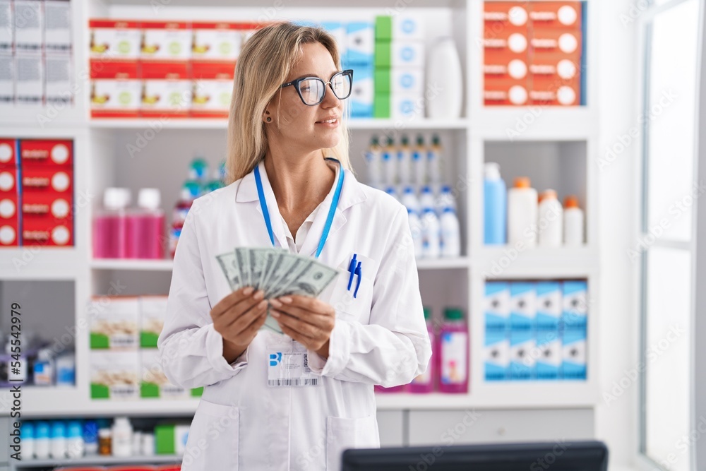 Young blonde woman pharmacist smiling confident counting dollars at pharmacy