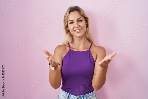 Young blonde woman standing over pink background smiling cheerful offering hands giving assistance and acceptance.