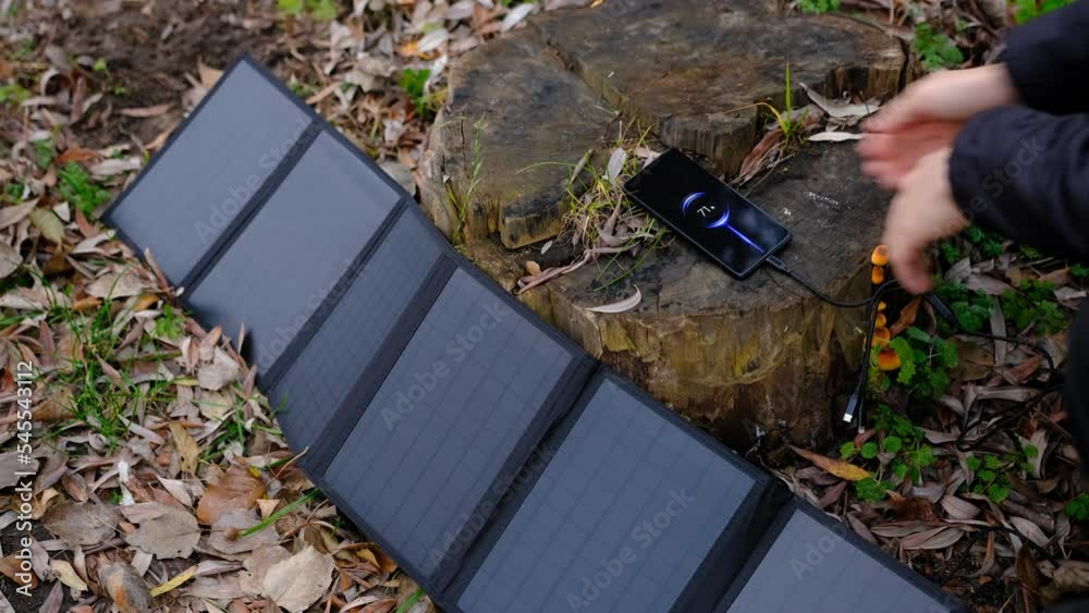 Hands plug in portable solar panel charging smartphone by mutlipurpose cable, power bank, watches, laptops nature background. Clean energy for using in camping or at home when NO electricity