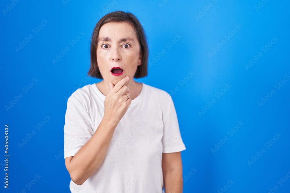 Middle age hispanic woman standing over blue background looking fascinated with disbelief, surprise and amazed expression with hands on chin Middle age hispanic woman standing over blue background looking fascinated with disbelief, surprise and amazed expression with hands on chin