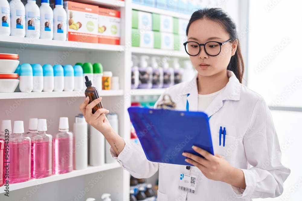 Young chinese woman pharmacist reading clipboard holding bottle at pharmacy Young chinese woman pharmacist reading clipboard holding bottle at pharmacy