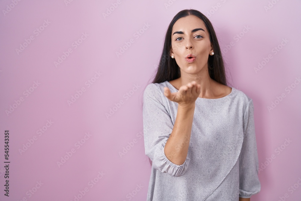 Young brunette woman standing over pink background looking at the camera blowing a kiss with hand on air being lovely and sexy. love expression. Young brunette woman standing over pink background looking at the camera blowing a kiss with hand on air being lovely and sexy. love expression.