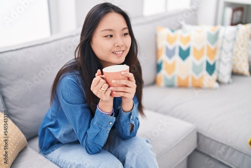 Young chinese woman drinking coffee sitting on sofa at home