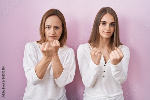 Middle age mother and young daughter standing over pink background ready to fight with fist defense gesture, angry and upset face, afraid of problem