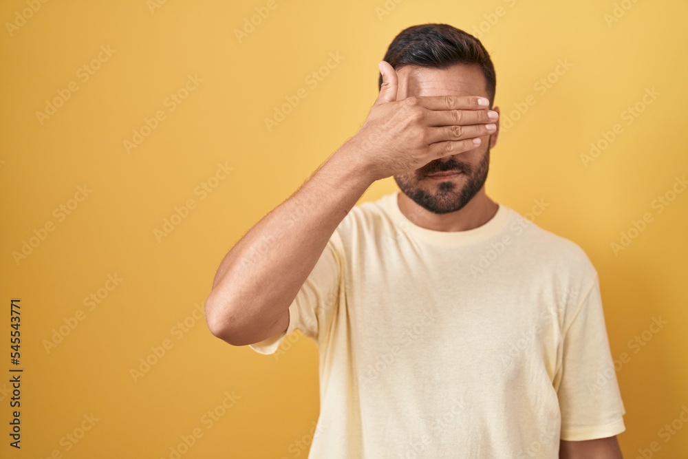 Handsome hispanic man standing over yellow background covering eyes with hand, looking serious and sad. sightless, hiding and rejection concept Handsome hispanic man standing over yellow background covering eyes with hand, looking serious and sad. sightless, hiding and rejection concept