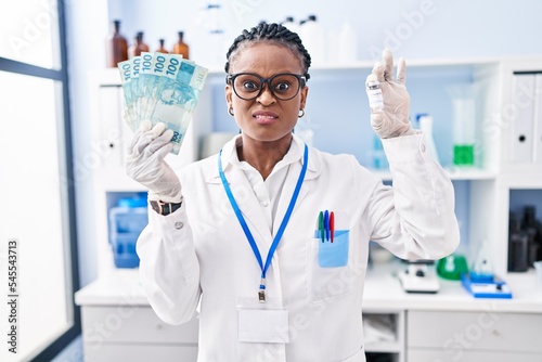 African woman with braids working at scientist laboratory holding money clueless and confused expression. doubt concept.