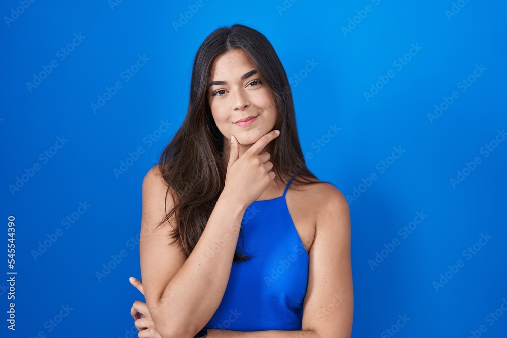 Hispanic woman standing over blue background looking confident at the camera smiling with crossed arms and hand raised on chin. thinking positive. Hispanic woman standing over blue background looking confident at the camera smiling with crossed arms and hand raised on chin. thinking positive.