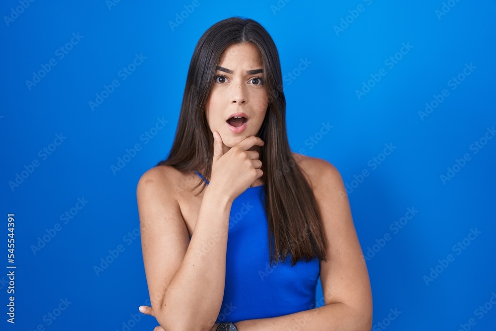 Hispanic woman standing over blue background looking fascinated with disbelief, surprise and amazed expression with hands on chin Hispanic woman standing over blue background looking fascinated with disbelief, surprise and amazed expression with hands on chin