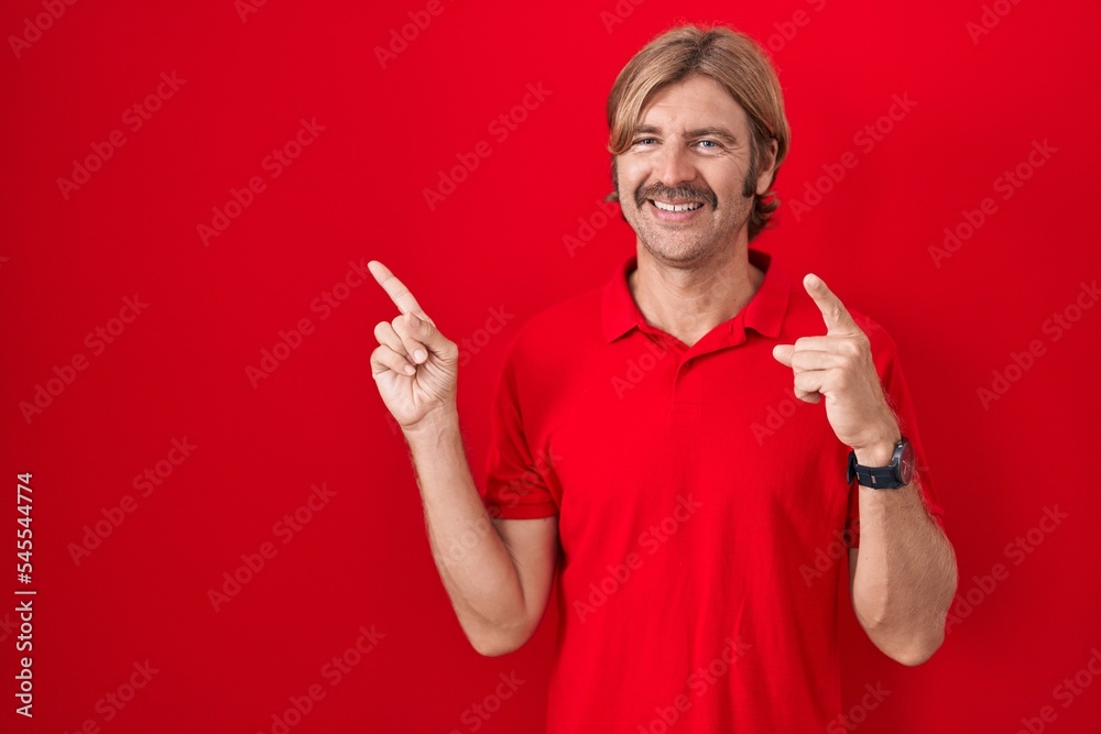 Caucasian man with mustache standing over red background smiling and looking at the camera pointing with two hands and fingers to the side.