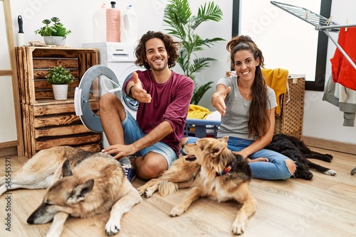 Young hispanic couple doing laundry with dogs smiling friendly offering handshake as greeting and welcoming. successful business.
