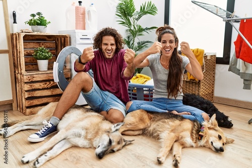 Young hispanic couple doing laundry with dogs angry and mad raising fists frustrated and furious while shouting with anger. rage and aggressive concept.