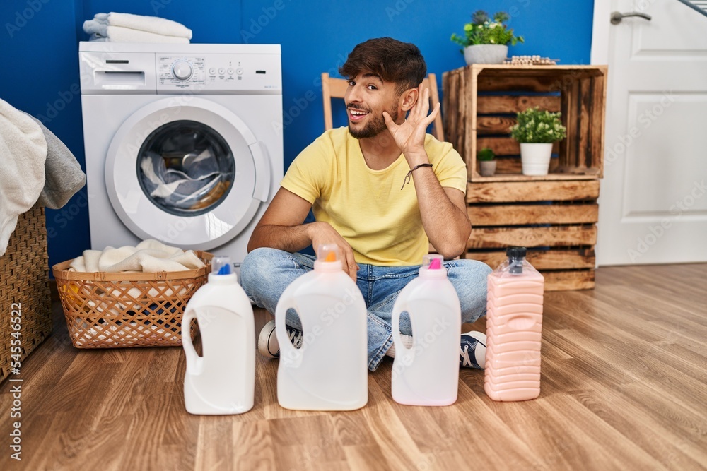 Arab man with beard doing laundry sitting on the floor with detergent bottle smiling with hand over ear listening and hearing to rumor or gossip. deafness concept. Arab man with beard doing laundry sitting on the floor with detergent bottle smiling with hand over ear listening and hearing to rumor or gossip. deafness concept.