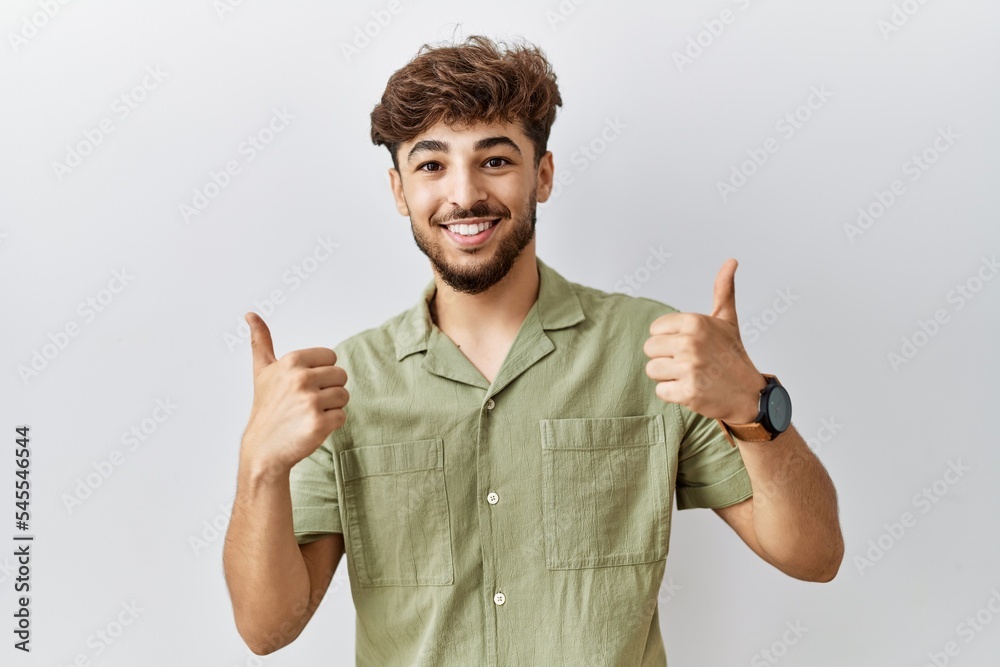 Young arab doctor man standing over isolated background success sign doing positive gesture with hand, thumbs up smiling and happy. cheerful expression and winner gesture. Young arab doctor man standing over isolated background success sign doing positive gesture with hand, thumbs up smiling and happy. cheerful expression and winner gesture.