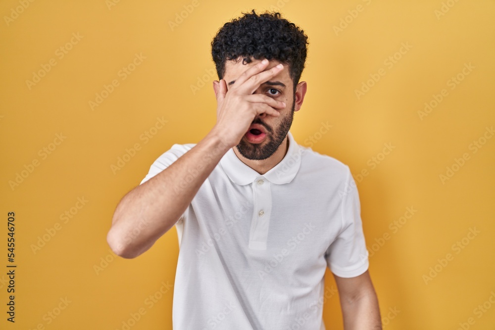 Arab man standing over yellow background peeking in shock covering face and eyes with hand, looking through fingers with embarrassed expression. Arab man standing over yellow background peeking in shock covering face and eyes with hand, looking through fingers with embarrassed expression.